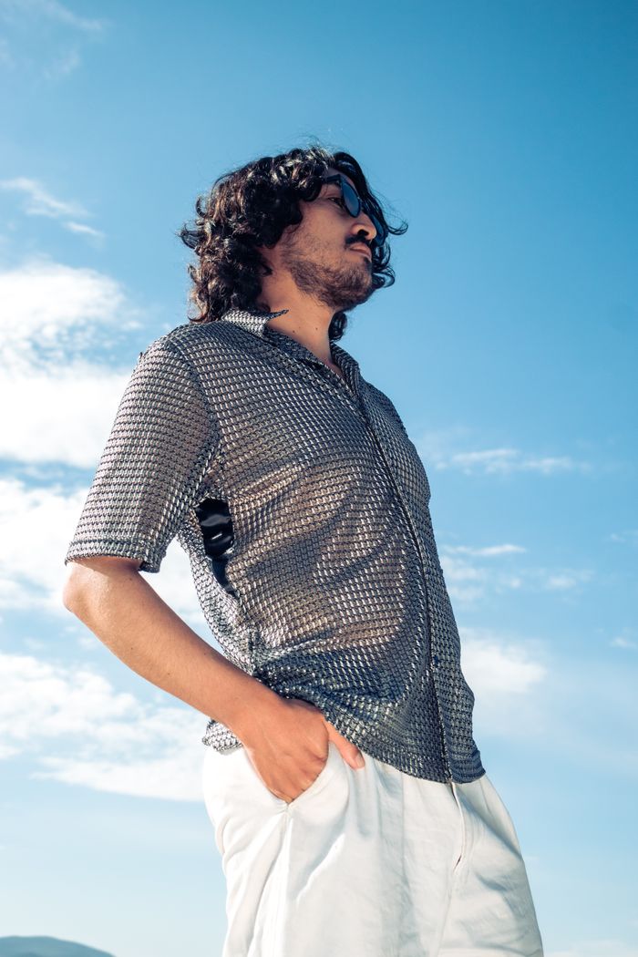 About A fashionable man with long hair and sunglasses, standing outdoors in a gray shirt and white pants.