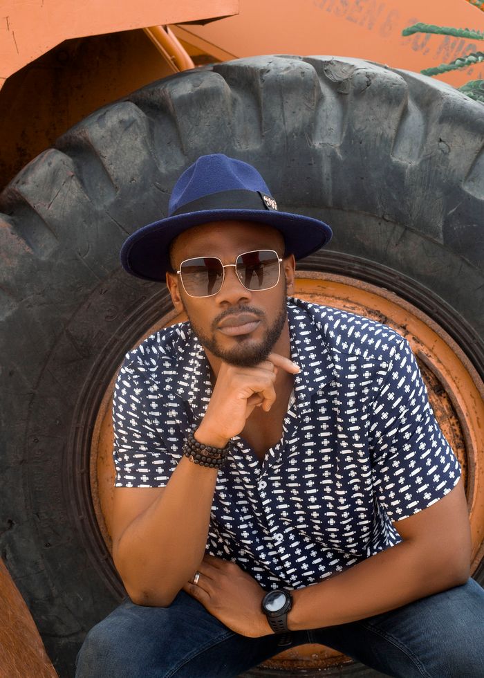 About Fashionable portrait of a stylish man in blue hat and sunglasses posing outdoors with tractor tire backdrop.