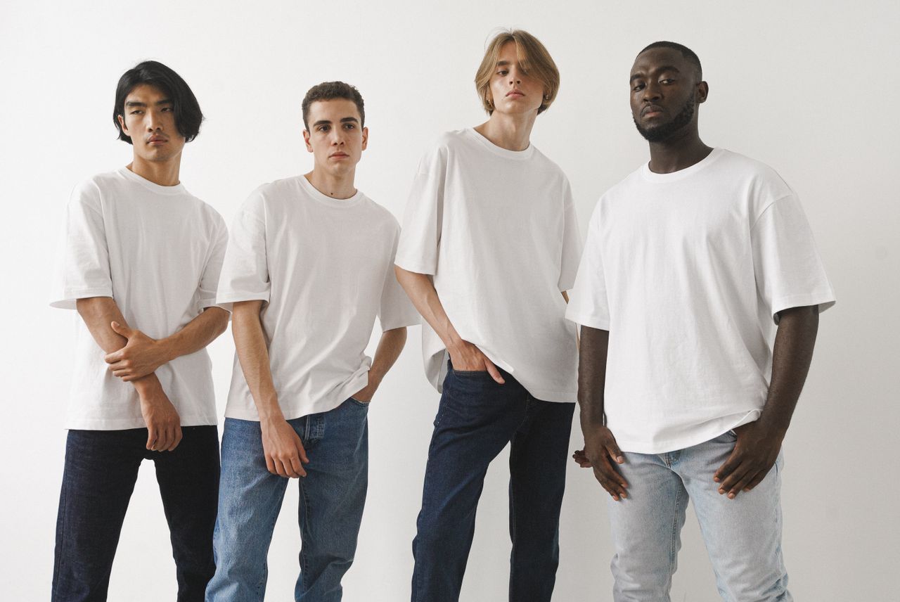Home A group of four diverse men modeling white t-shirts and jeans in a studio setting.
